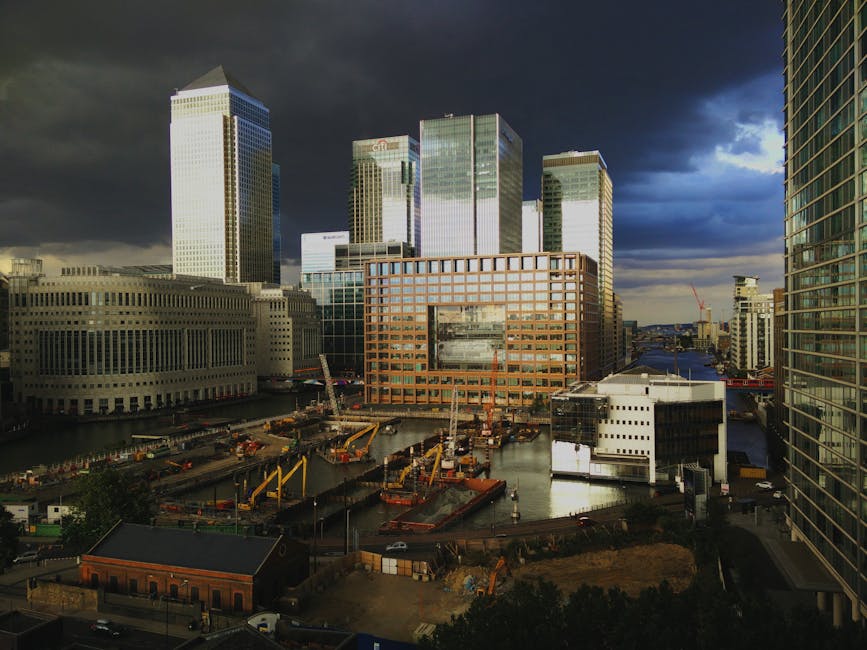 A daytime view of a busy urban construction site surrounded by tall modern office buildings with glass and steel facades in the Docklands area of London, indicating ongoing building or renovation work