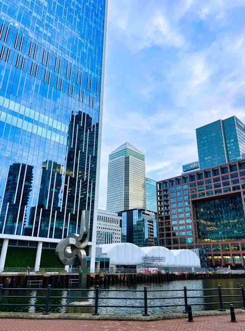 A daytime view of a busy urban construction site surrounded by tall modern office buildings with glass and steel facades in the Docklands area of London, indicating ongoing building or renovation work