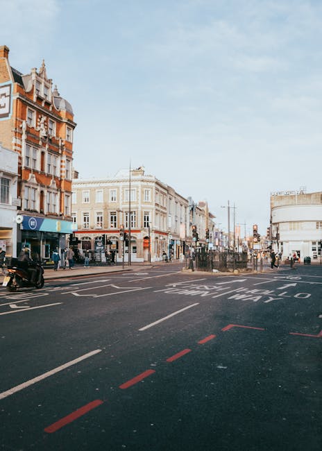 A busy street scene in Camden Town NW1, London, with pedestrians crossing at a traffic light and storefronts lining the pavement. An elevated railway bridge, painted with the words 'CAMDEN LOCK' in la